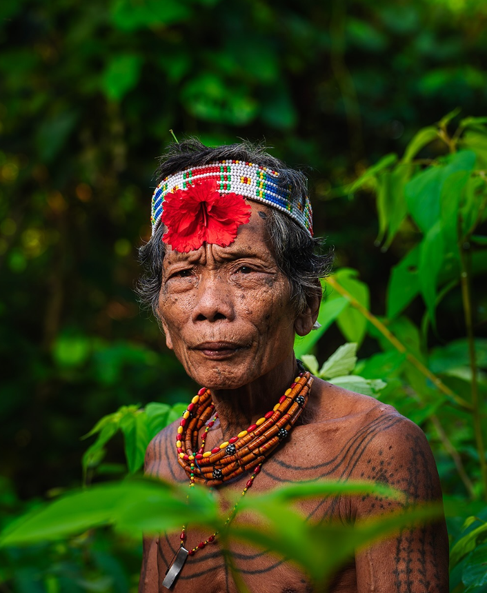 Uomo indigeno anziano con tatuaggi facciali, fascia colorata con perline e un fiore rosso e collane di perline stratificate, circondato da fogliame verde.